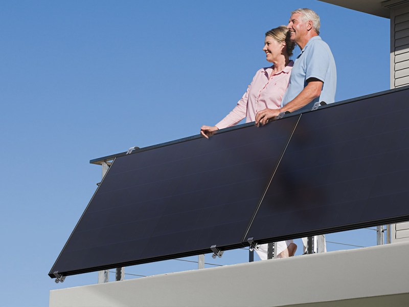 Zwei Personen stehen auf einem Balkon vor sonnigem Himmel. Am Geländer ist ein Balkonkraftwerk angebracht.