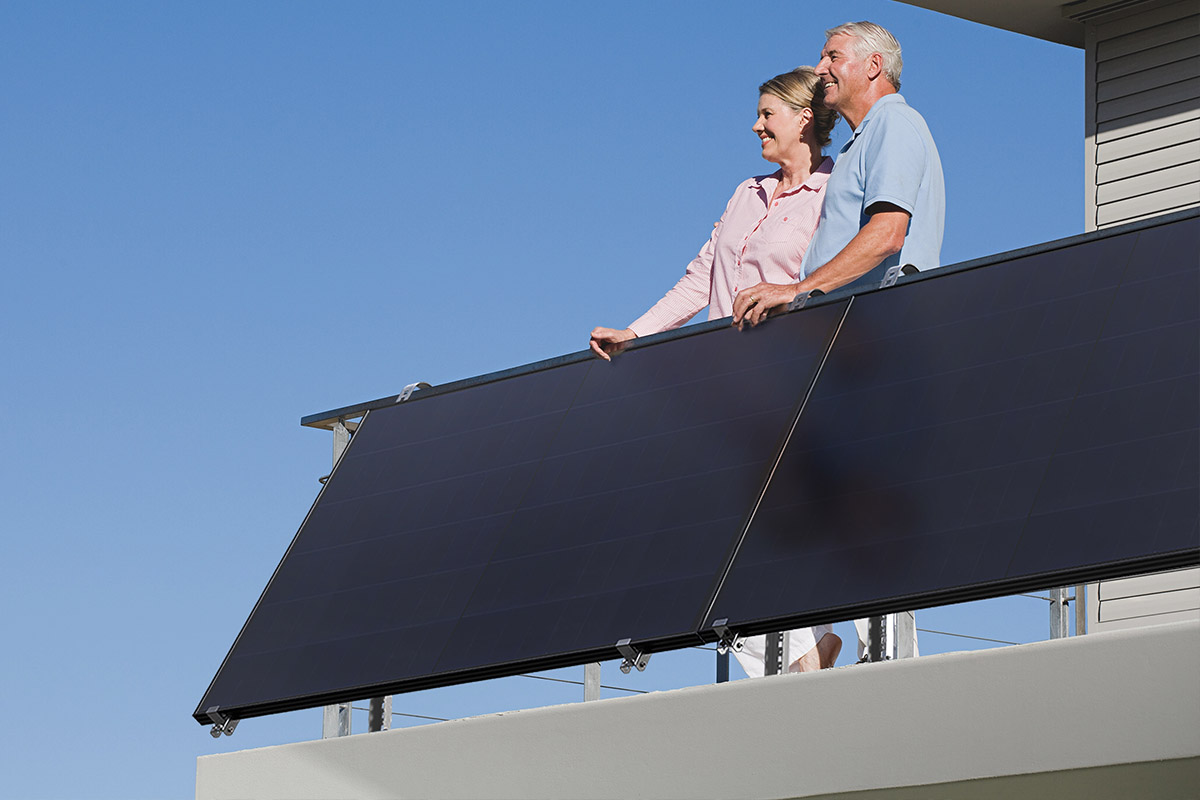 Zwei Personen stehen auf einem Balkon vor sonnigem Himmel. Am Geländer ist ein Balkonkraftwerk angebracht.