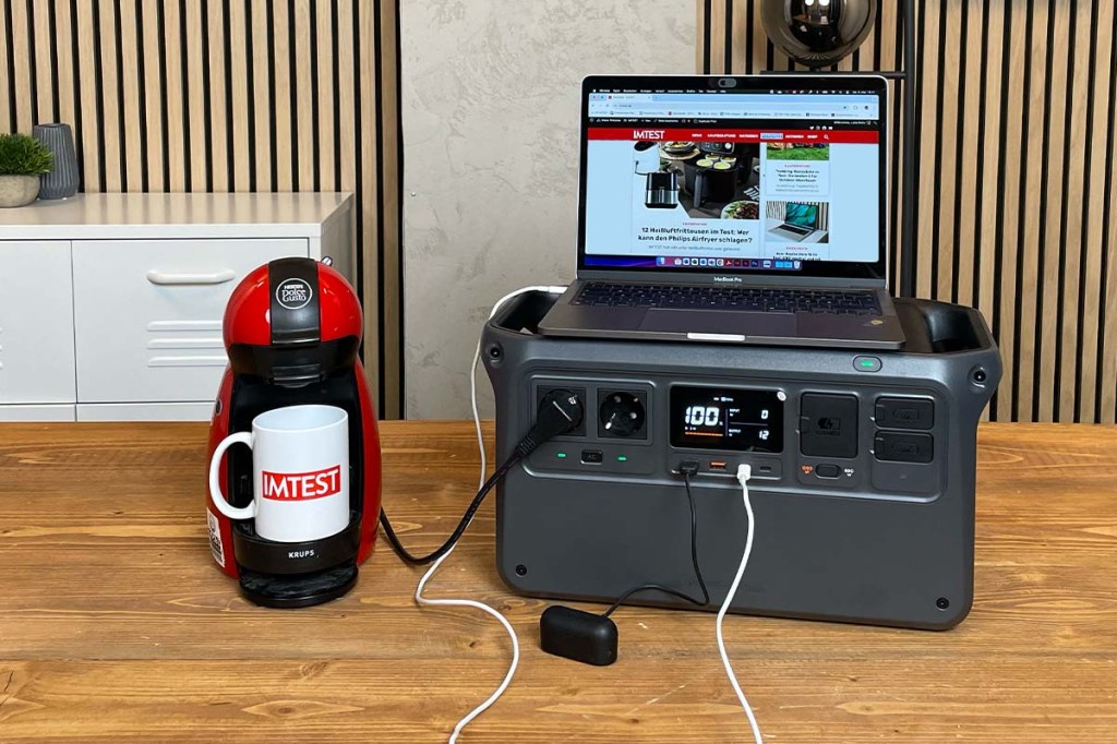 Die DJI-Powerstation auf einem Holztisch in wohnlicher Umgebung. Verschiedene Geräte sind angeschlossen: Ein Laptop, Bluetooth-Kopfhörer und eine rote Kaffee-Pad-Maschine. Auf dem weißen Becher ist das IMTEST-Logo aufgedruckt.