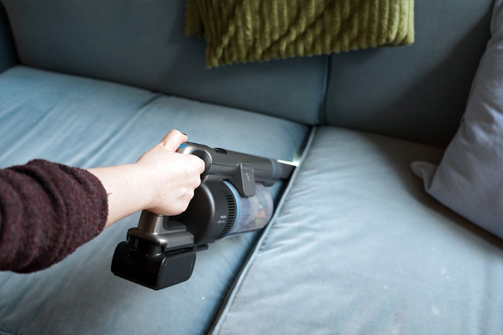 Eine Person saugt die Sofaritzen eines blauen Sofas mit dem Philips-Akkusauger und einem Fugen-Aufsatz.
