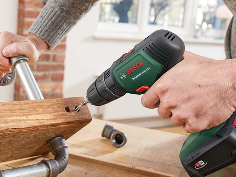 Hand hält ein Holzstück mit Rohren fest, eine andere einen Bohrer