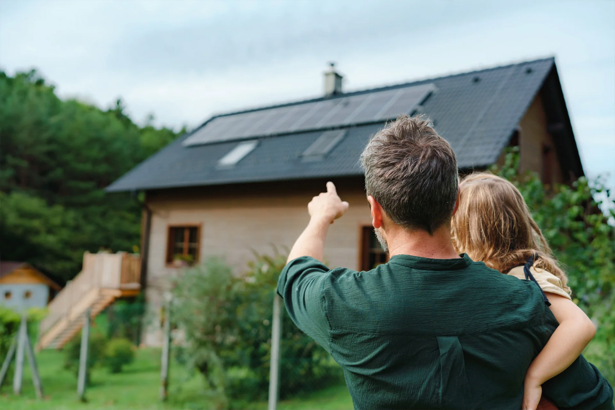 Ein Mann mit Kind auf dem Arm steht mit dem Rücken zur Kamera und zeigt auf ein Haus mit Solarpanels auf dem Dach.