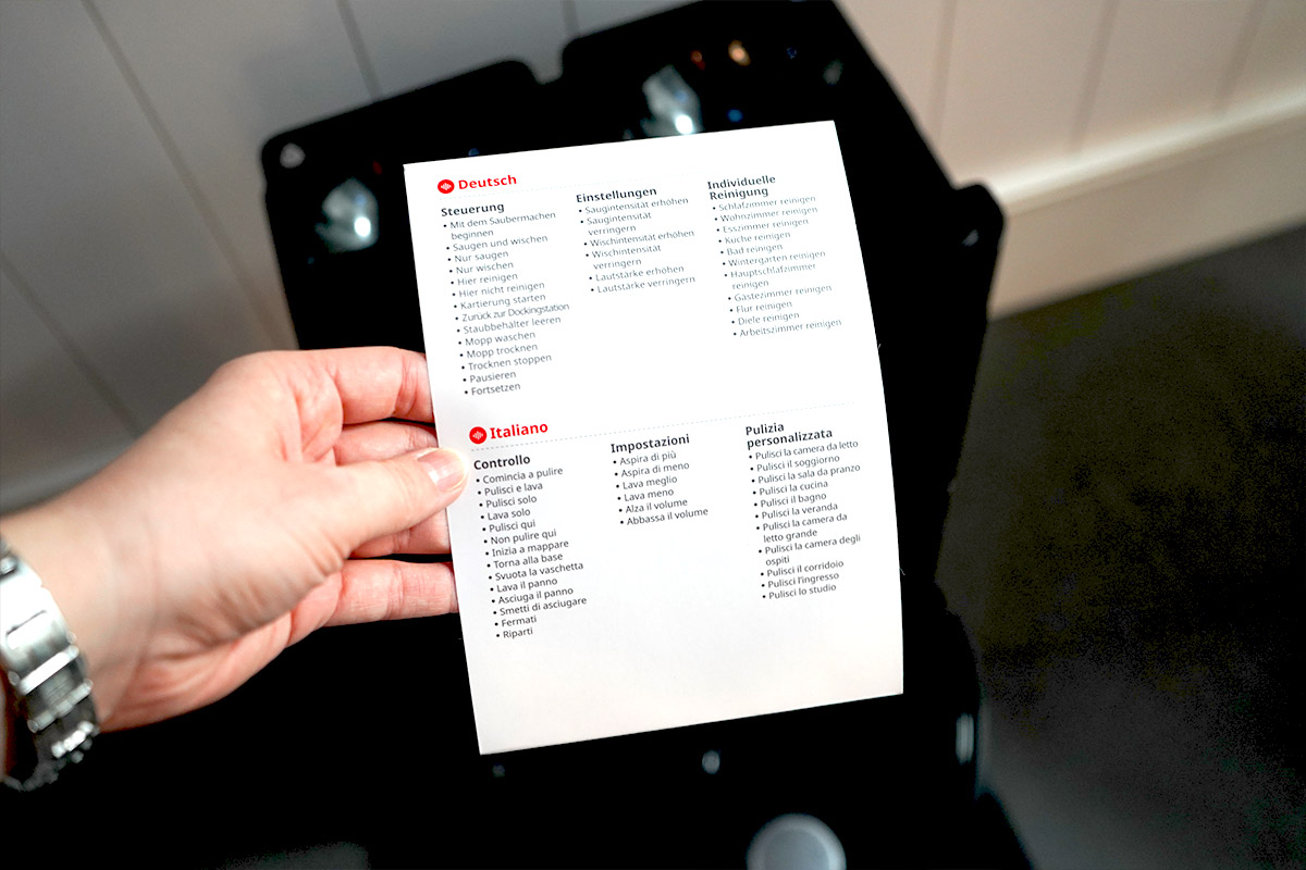 Eine Hand hält eine Liste in die Kamera. Im Hintergrund ist eine Saugroboter-Station zu sehen.