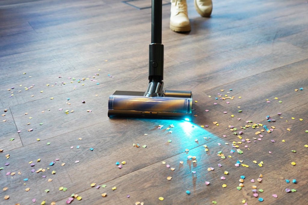 Eine Person saugt Konfetti von einem Holzfußboden mit dem Dreame Z40 Station. Ein türkis-blaues Licht ist an der Bodendüse zu sehen.