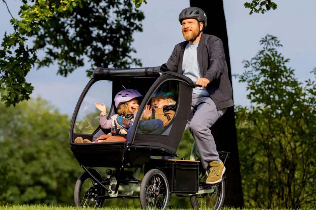 Mann mit einem Cargo-E-bike im Park, in der Transportkabine sitzen zwei Kinder