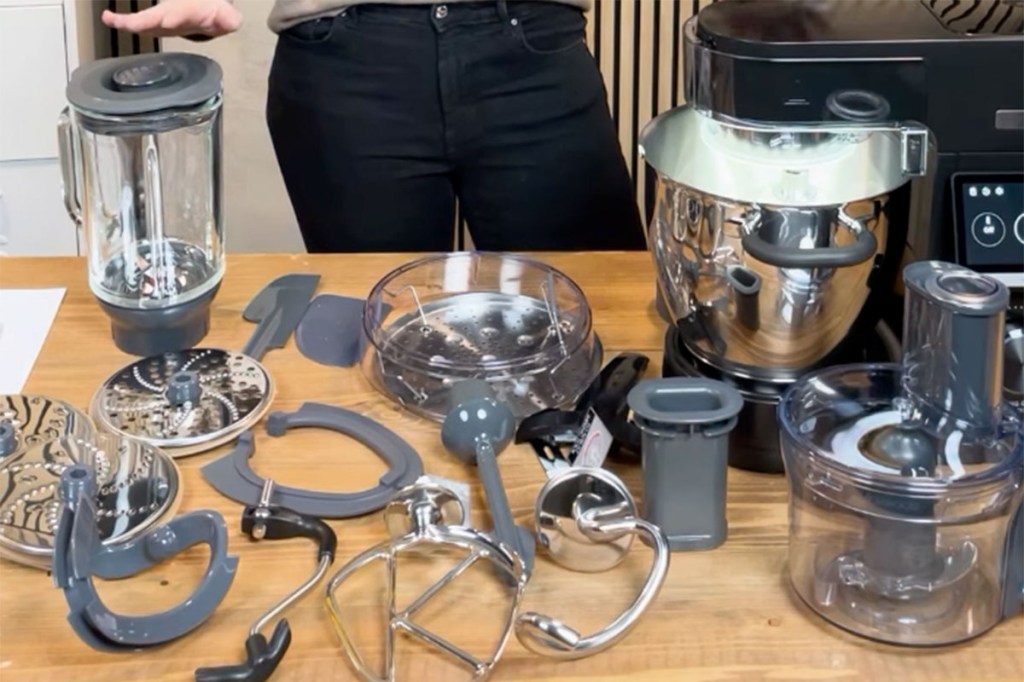 Der Kenwood Cooking Chef Sallys Edition mit mitgeliefertem Zubehör auf einem Holztisch. Dahinter steht eine Person.
