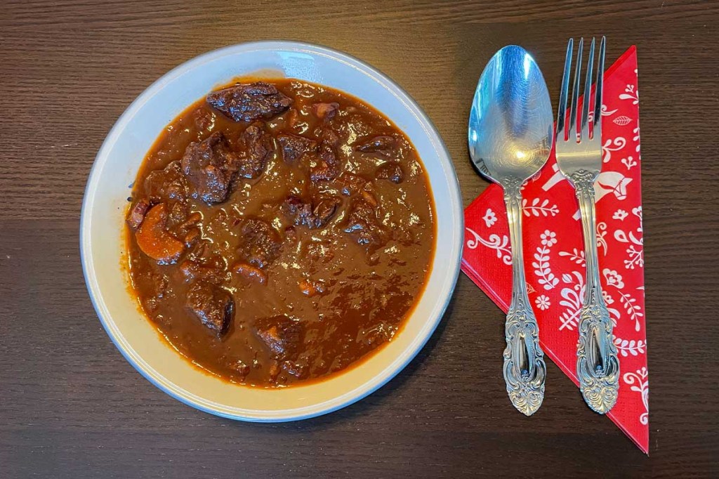 Ein Teller mit Gulasch auf einem Holztisch. Daneben: Gabel, Löffel und Serviette.