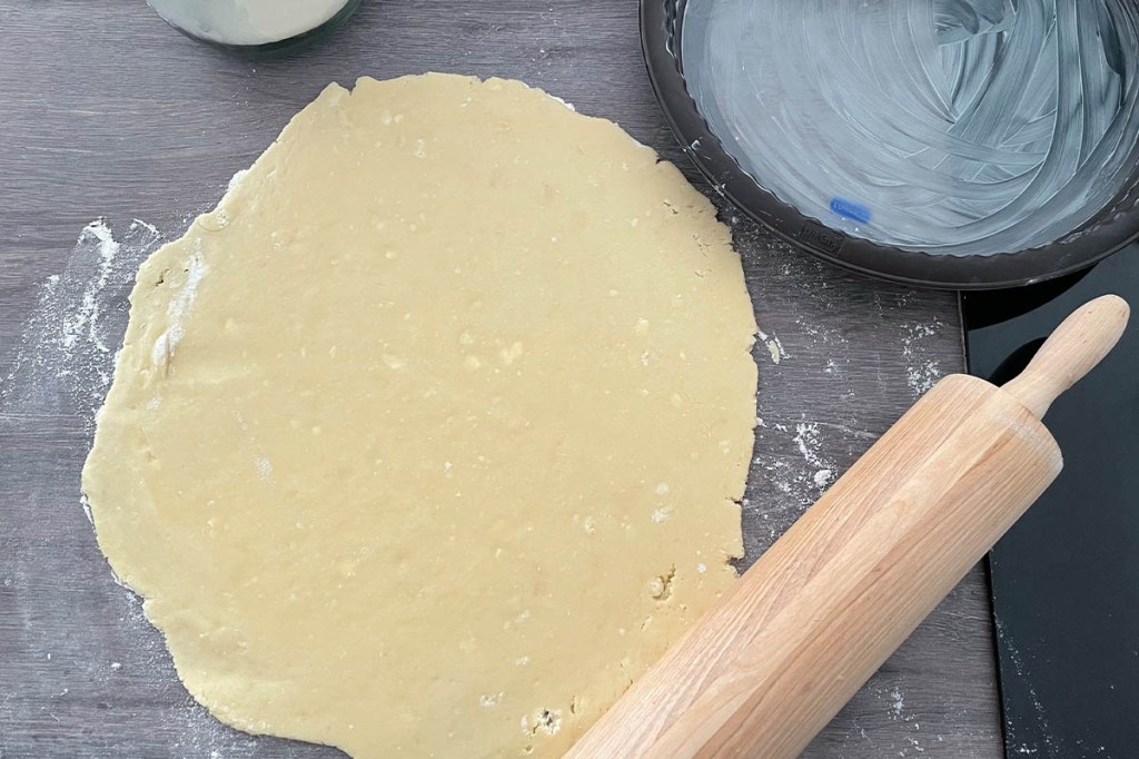 Ausgerollter Mürbeteig auf einer Arbeitsplatte. Daneben eine Kuchenform, ein Nudelholz und ein Mehlbehälter.