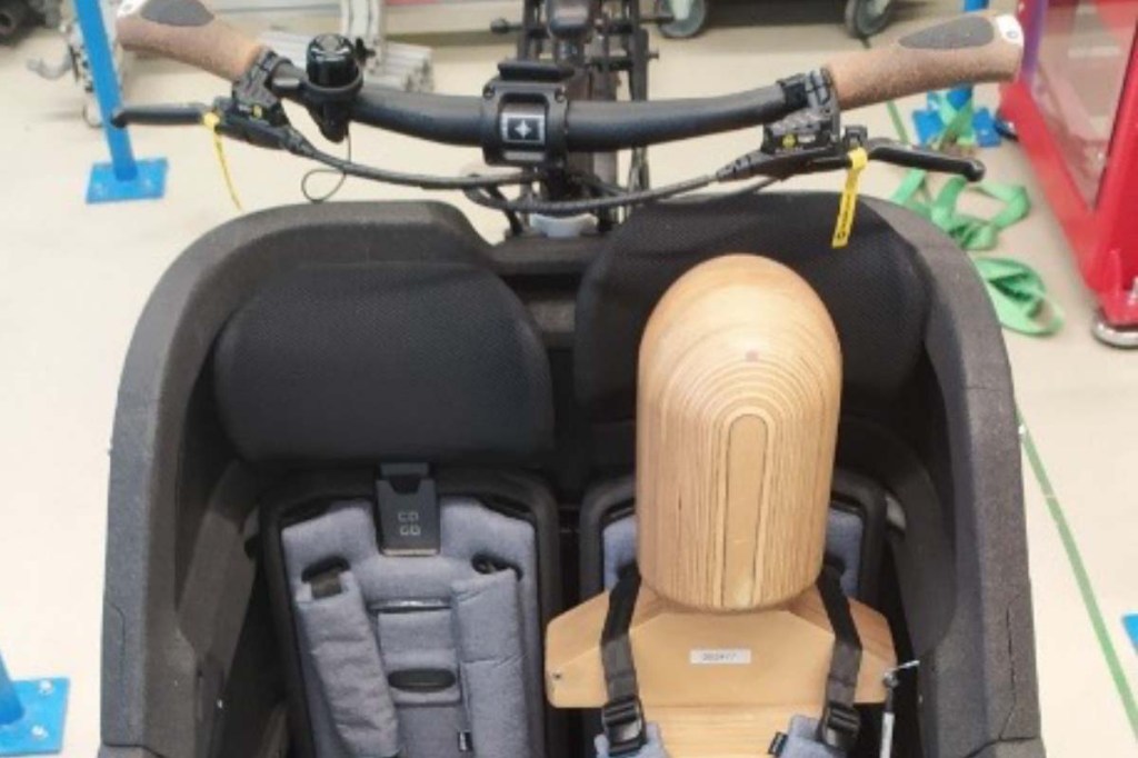 Holzpuppe sitzt in der Transportwanne eines Cargo-E-Bikes, dabei wird geprüft, ob der Kopf gegen den Lenker stößt