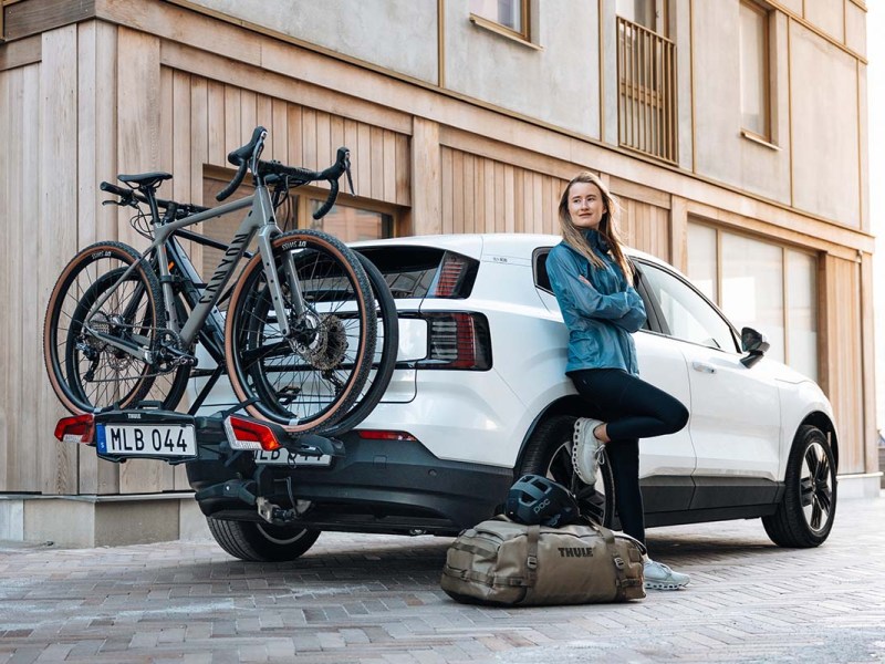 Frau steht vor einem weißen Auto, an diesem ist ein Heckträger mit zwei Gravelbikes befestigt
