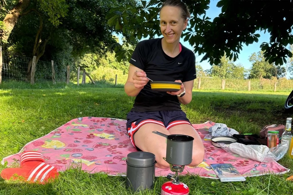 Frau sitzt auf einer Picknickdecke, isst von Campinggeschirr, davor eine Campingkocher