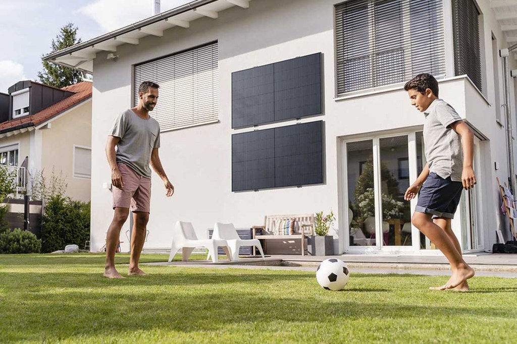 Vater und Sohn spielen Fußball vor einem weißen Haus mit Solarpanels an der Fassade.
