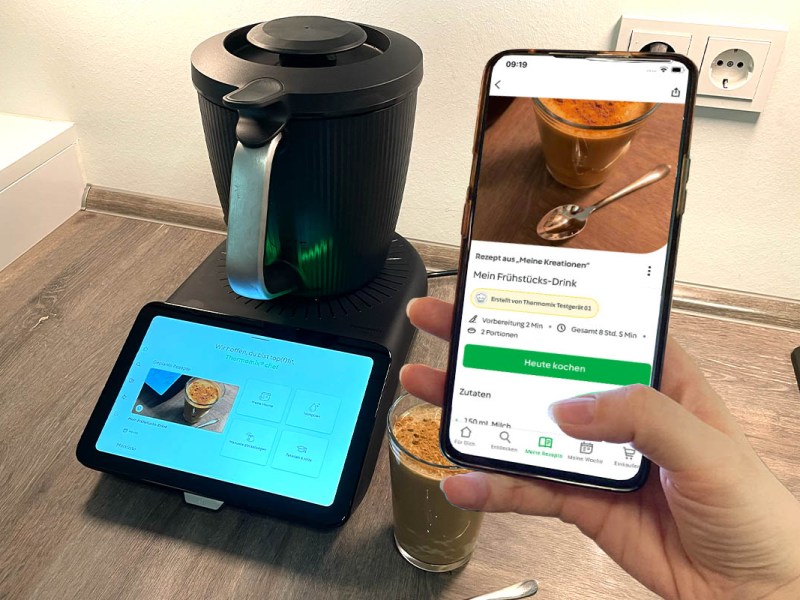 Eine Hand hält ein Smartphone mit Cookidoo-App in die Kamera. Im Hintergrund der Thermomix TM7.