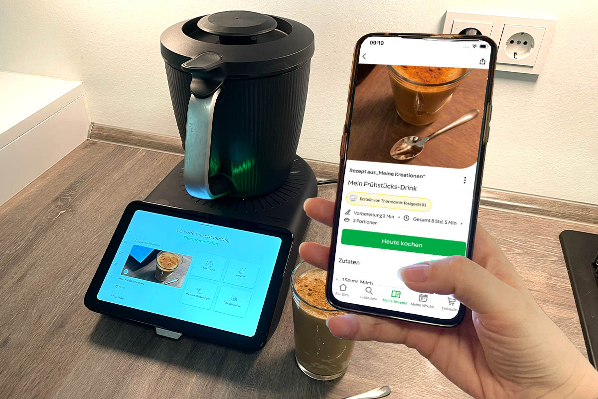 Eine Hand hält ein Smartphone mit Cookidoo-App in die Kamera. Im Hintergrund der Thermomix TM7.
