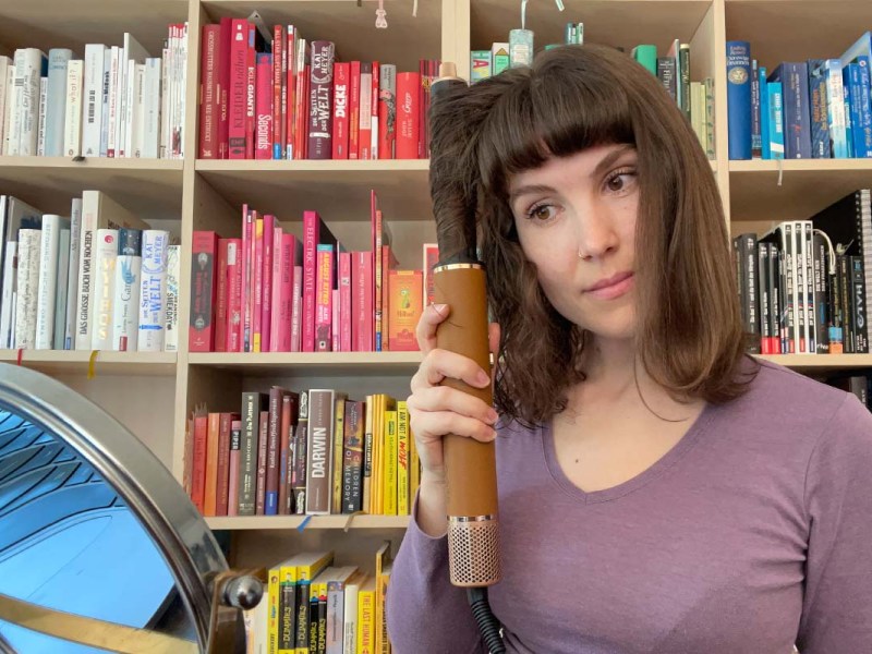 Eine Frau sitzt vor einem bunten Bücherregal und lockt sich mit dem Dreame Airstyle Pro und dem Lockenstab-Aufsatz die Haare.