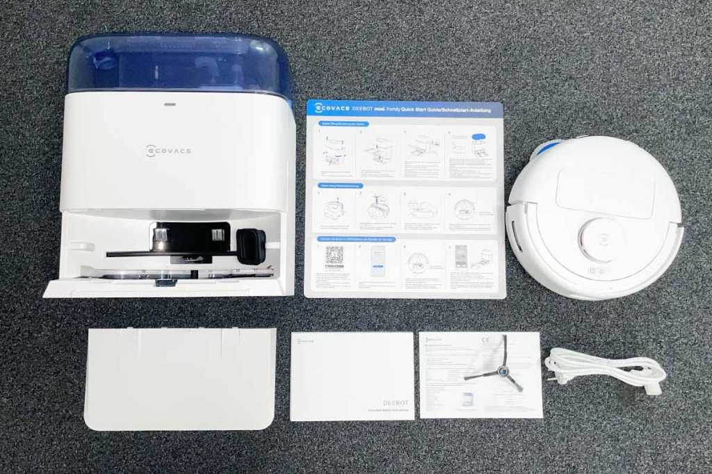 Der Ecovacs Deebot mini neben Station und Lieferzubehör auf einem dunklen Teppich