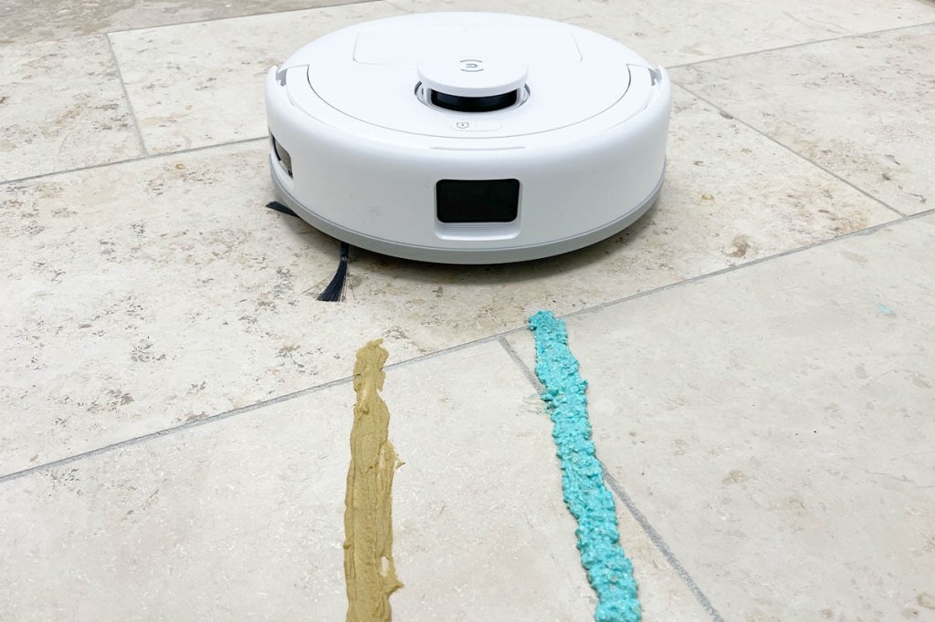 Der Deebot mini auf hellem Steinfußboden mit zwei bunten Schmutz-Streifen in Richtung Kamera.