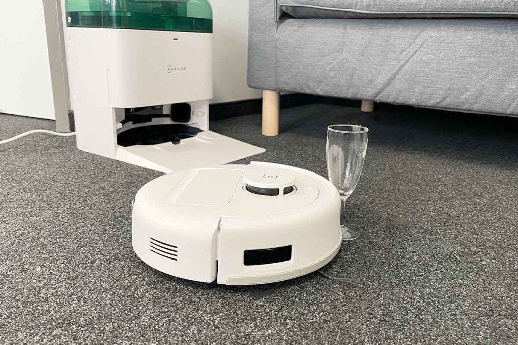 Der Deebot mini fährt gegen ein Trinkglas. Im Hintergrund die Station und ein Sofa.