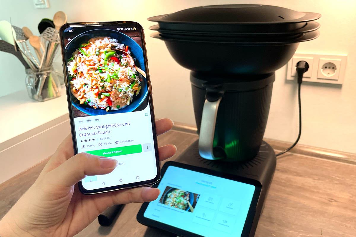 Eine Hand hält ein Smartphone mit Cookidoo-App in die Kamera. Dahinter der Thermomix TM7 auf einer Küchenarbeitsplatte.