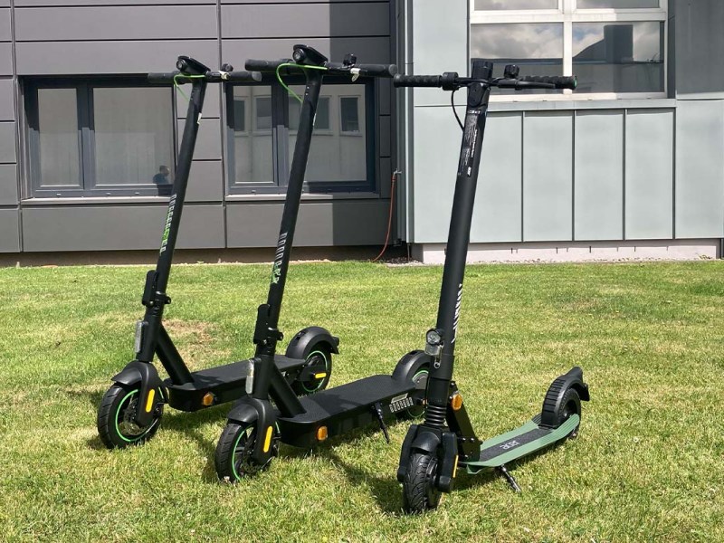 drei E-Scooter vor einem Firmengebäude auf einer Wiese stehend