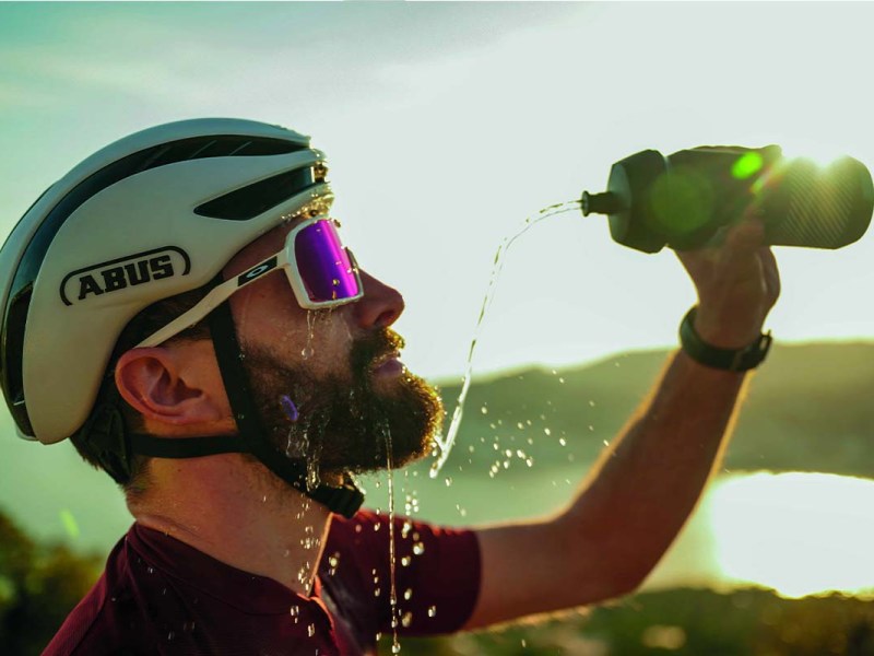 Mann mit Fahrradhelm und Sonnenbrille, bespritzt sich mit Wasser aus seiner Trinkflasche, Sonne im HIntergrund