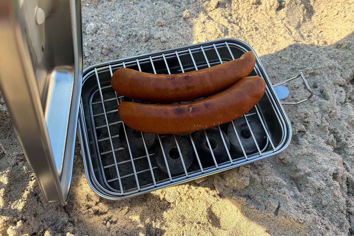 Zwei Würstchen auf einem kompakten Grill liegend, der Grill steht im Sand