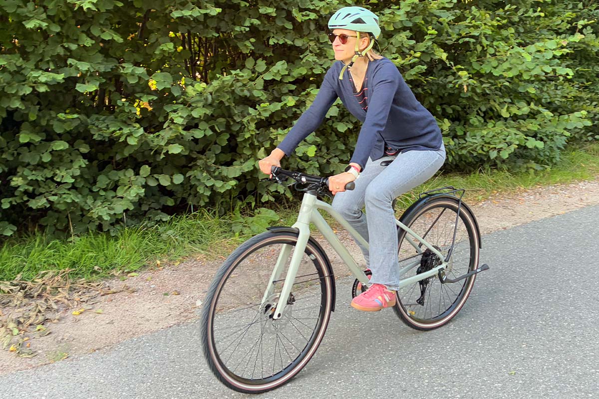 Frau fährt mit einem Fahrrad eine STraße entlang, Büsche im Hintergrund