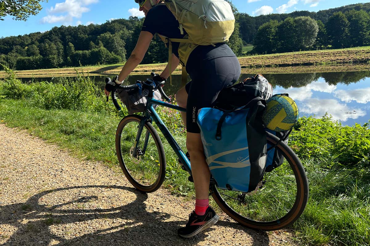 Anschnitt eines fahrradfahrers von hinten, der gepäckträger ist mit taschen voll beladen, ein kanal und natur im HIntergrund