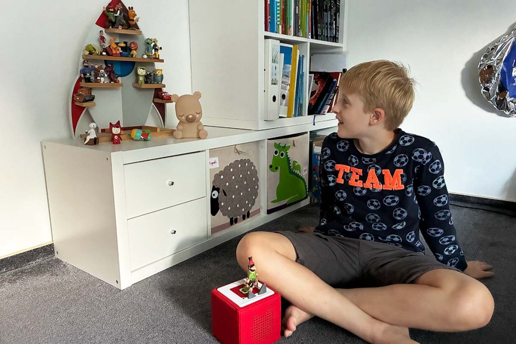 Kind sitzt in seinem Zimmer, vor ihm eine rote Tonie-Box, heben ihm ein Regal mit vielen Tonie-Figuren