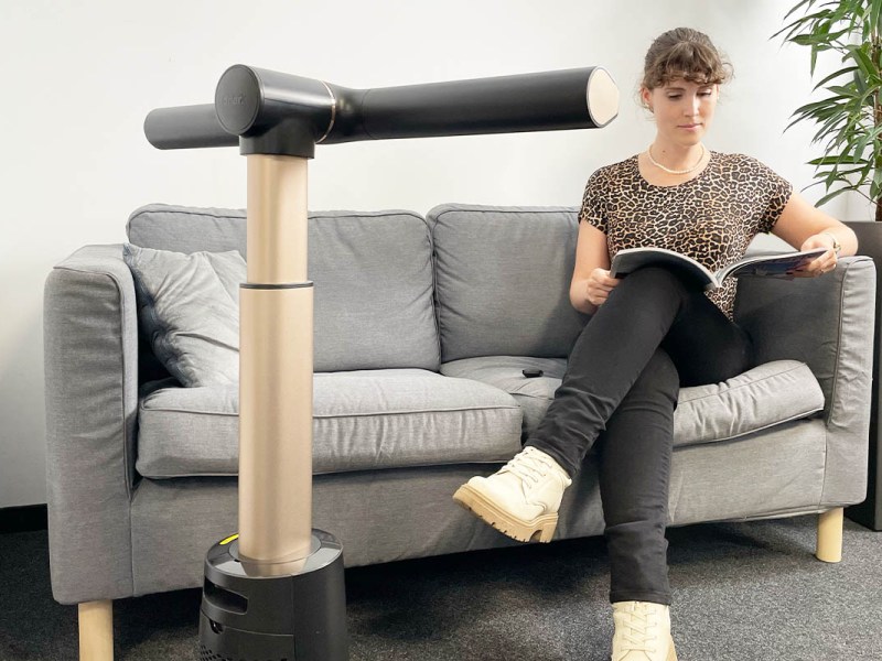 Eine Frau sitzt im T-Shirt auf einem Sofa und liest. Davor der Shark TurboBlade Ventilator.