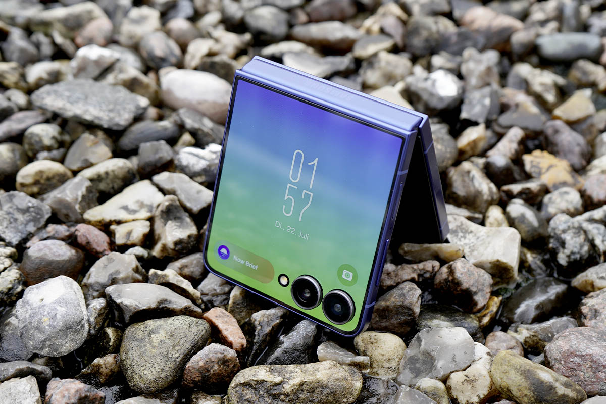 Das Samsung Galaxy Z Flip7 steht halb aufgeklappt mit eingeschaltetem Außendisplay auf einer Kiesoberfläche.