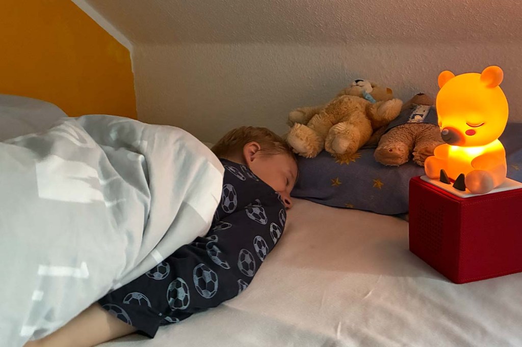 kind liegt in einem Bett und schläft, neben ihm steht eine toniebox mit einem leuchtenden Bär obendrauf