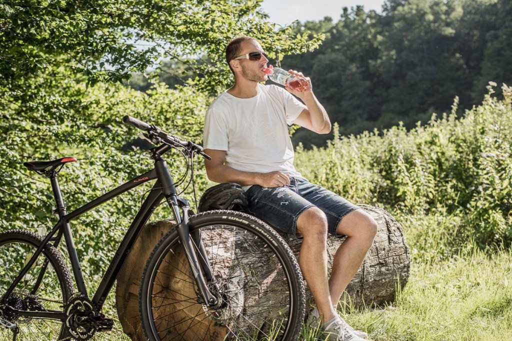 Mann auf einem Baumstamm sitzend, er trinkt aus einer Wasserflasche, sein FAhrrad steht neben ihm, Bäume und STräucher im Hintergrund